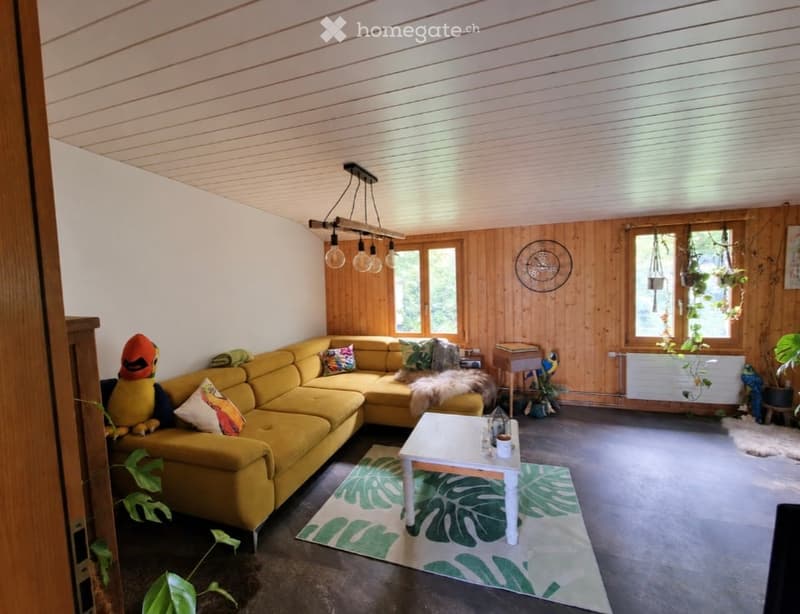 Bright living room with wooden panel walls, large windows, yellow sectional sofa, white coffee table, and decorative plants.
