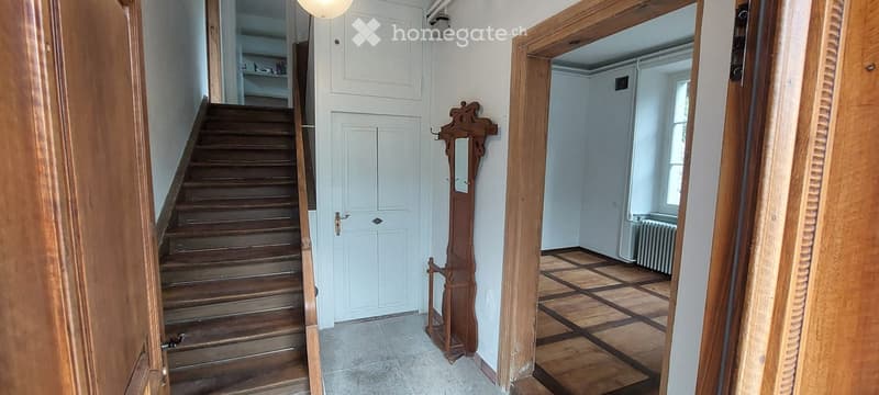 Hallway with wooden staircase, white double doors, and a coat rack. The adjacent room has a checkered wooden floor and a radiator.