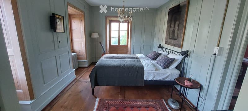 Cozy bedroom with wooden floors, a double bed with grey and white bedding, a nightstand with a lamp, a small table with a pink duck figurine, a guitar leaning against the wall, and a chandelier hanging from the ceiling.