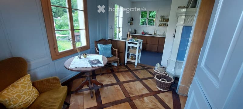 A cozy, well-lit room with a mix of living and kitchen areas. Features a patterned sofa, a round wooden table with a vase and book, a wooden chair with a cushion, and a small kitchen area with cabinets and a counter.
