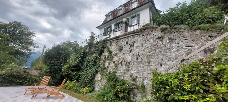 A raised outdoor area with wooden chairs, a stone retaining wall, and a house with a balcony above.