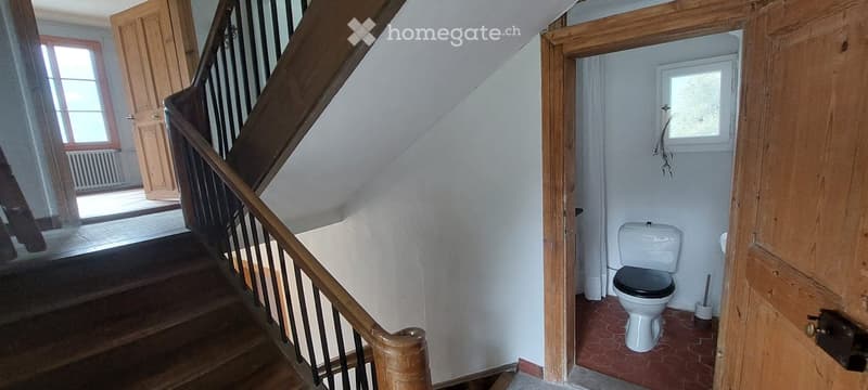 A narrow hallway with wooden stairs and a small adjacent bathroom featuring a toilet and a window.