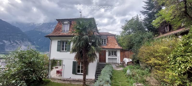 A two-story white house with green shutters and a red-tiled roof, situated in a lush garden with a palm tree, overlooking a mountainous landscape.