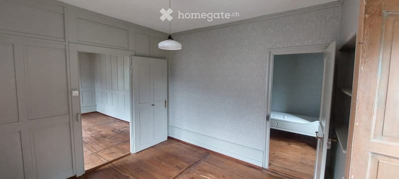Empty room with wooden flooring, light grey walls, and a hanging light fixture. Adjacent rooms are also empty with similar decor.