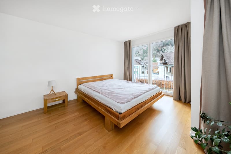 Bright bedroom with wooden flooring, a wooden bed with white bedding, a nightstand with a lamp, and large windows with curtains offering a view of the neighborhood.