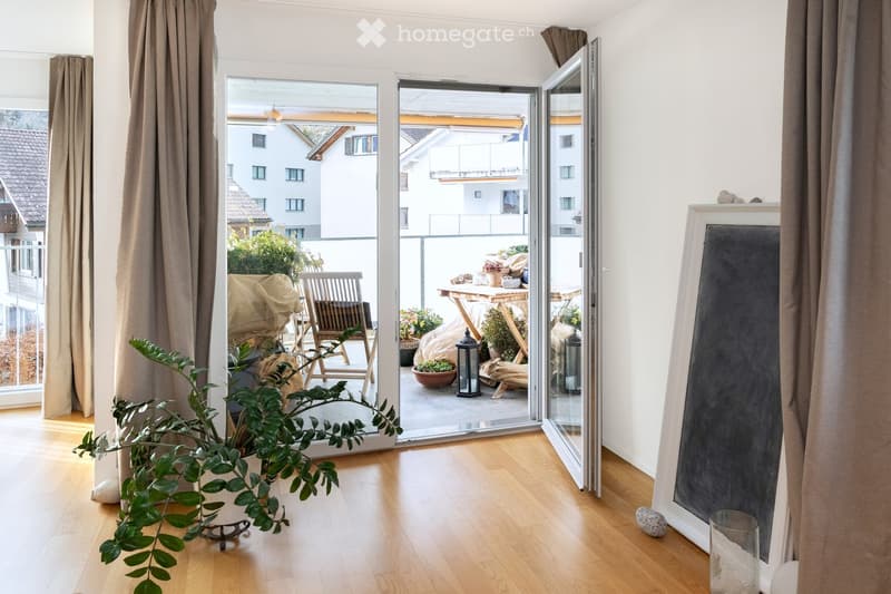Bright living space with wooden flooring, large sliding glass doors opening to a balcony, potted plants, a chair, a small table, and a mirror on the wall.