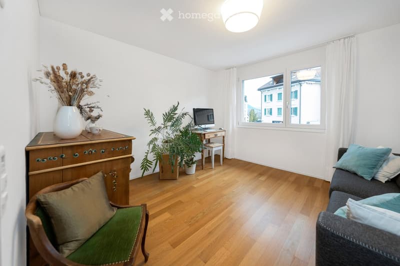 A well-lit living room with wooden flooring, a grey sofa with blue pillows, a wooden armchair with green cushions, a wooden side table with a vase of dried flowers, a small desk with a computer, and a potted plant near the window.
