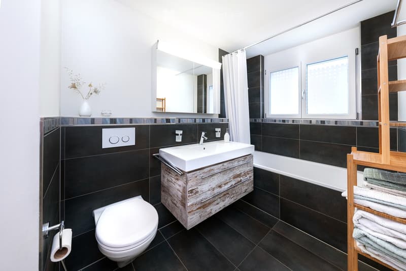 Modern bathroom with dark tiled walls and floor, white sink, toilet, bathtub, and a wooden towel rack with folded towels. A large mirror above the sink and a vase with dried flowers on the windowsill.