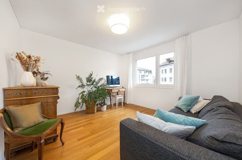Bright living room with wooden flooring, large windows, a grey sofa with blue pillows, a wooden armchair, a small table with a computer, a decorative vase with dried flowers, and a potted plant.