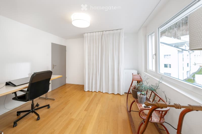 Bright, modern room with wooden flooring, large windows, a desk with a laptop, a comfortable chair, and a small wooden shelf with potted plants.