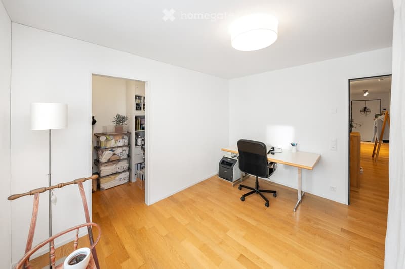 A well-lit, minimalist home office with a wooden floor, white walls, and a large ceiling light. The room features a modern desk with a chair, a printer, and a floor lamp. A small shelf with storage bins and a potted plant is visible in the background.