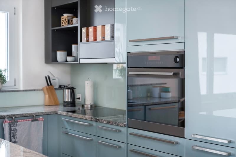 Modern kitchen with light blue cabinets, granite countertop, built-in oven, knife block, coffee maker, and various kitchen items on shelves and counter.
