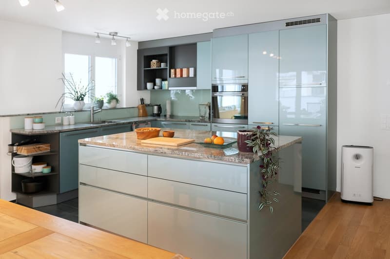 Modern kitchen with grey and white cabinets, a central island with a marble countertop, and various appliances including a refrigerator, oven, and sink. The island features a cutting board, bowls, and a potted plant. A white air purifier is placed on the