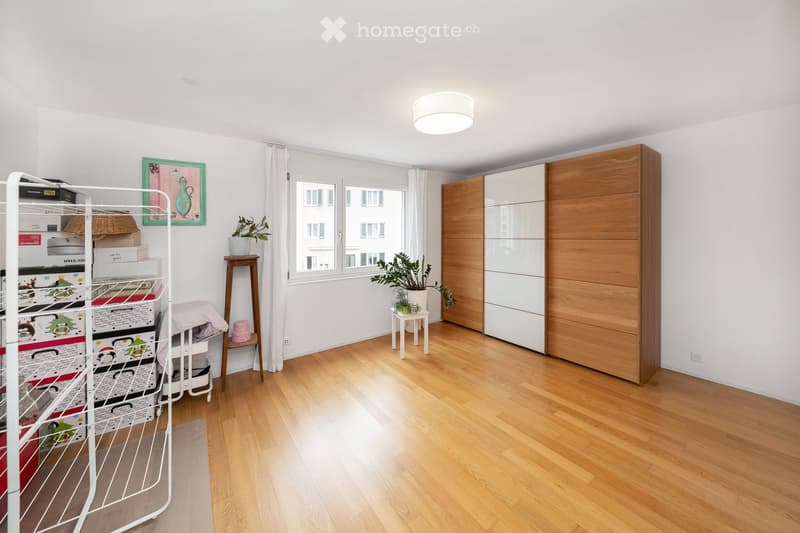 A well-lit room with wooden flooring, a large window with white curtains, a wardrobe, a small table with a potted plant, a storage rack with boxes, and a picture frame on the wall.