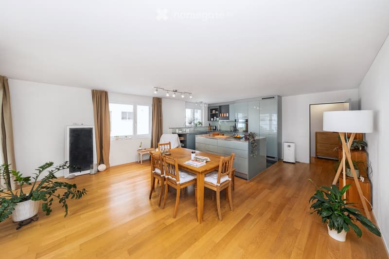 Bright, open-plan living and dining area with wooden flooring, modern kitchen, dining table with chairs, floor lamp, and potted plants.