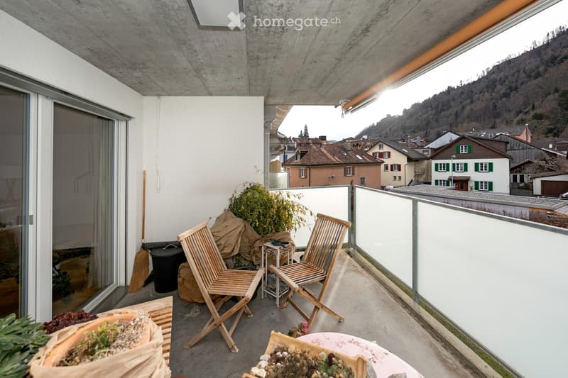Balcony with modern design, featuring two wooden chairs, a small table, and potted plants, overlooking a town with traditional houses and a hilly landscape.