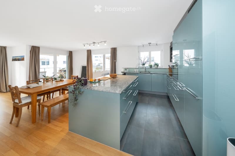 Modern kitchen with light blue cabinets, granite countertop, wooden dining table with chairs, and large windows with curtains.