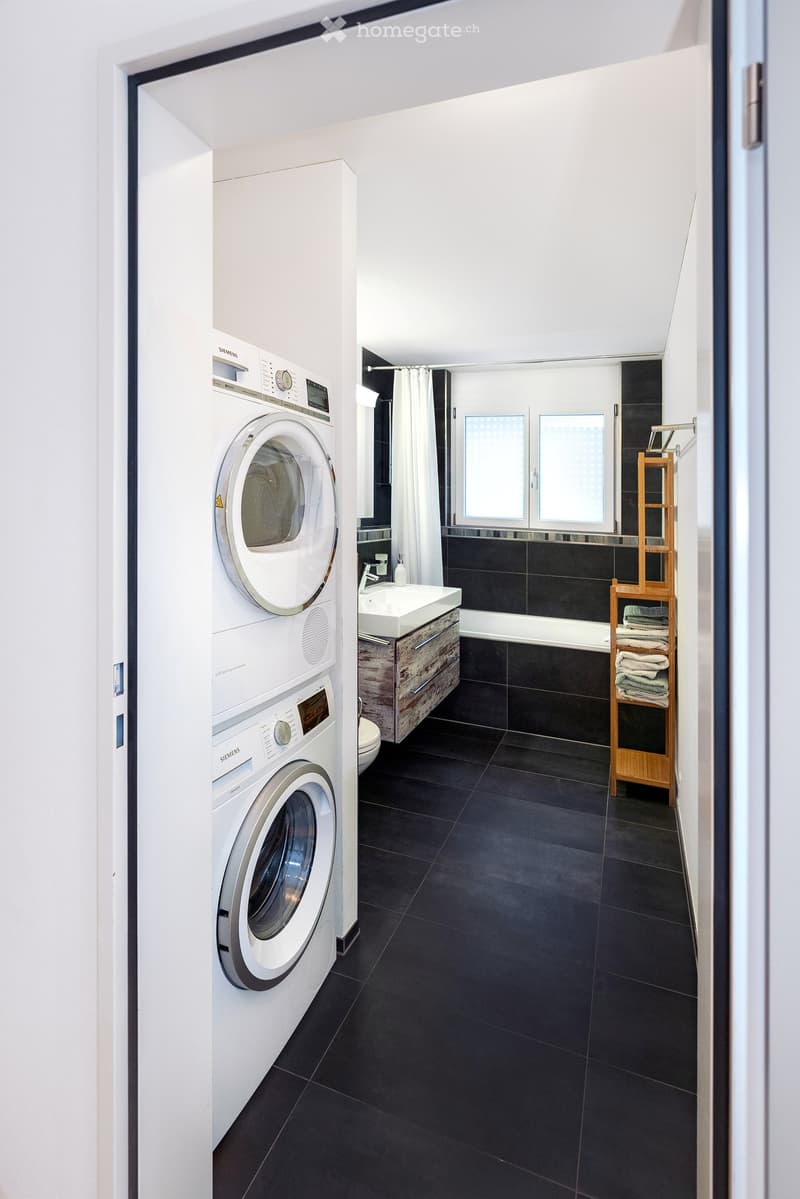 Modern bathroom with black tiled floor, white washing machine, sink with wooden cabinet, bathtub, and towel rack.