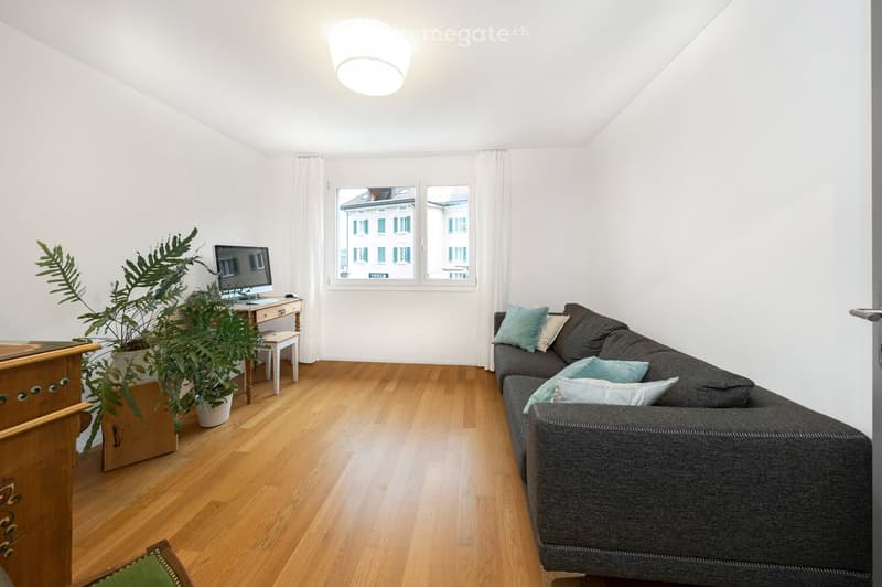 Living room with wooden flooring, large window with white curtains, grey sofa with light blue pillows, small table with a screen, and a potted plant.