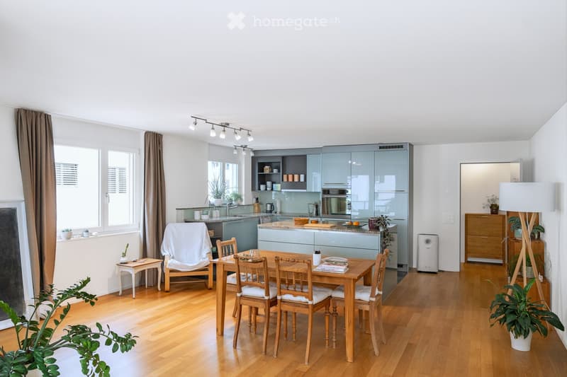 Bright, open-plan living and dining area with wooden flooring, modern kitchen cabinets, dining table with chairs, and various decorative plants.