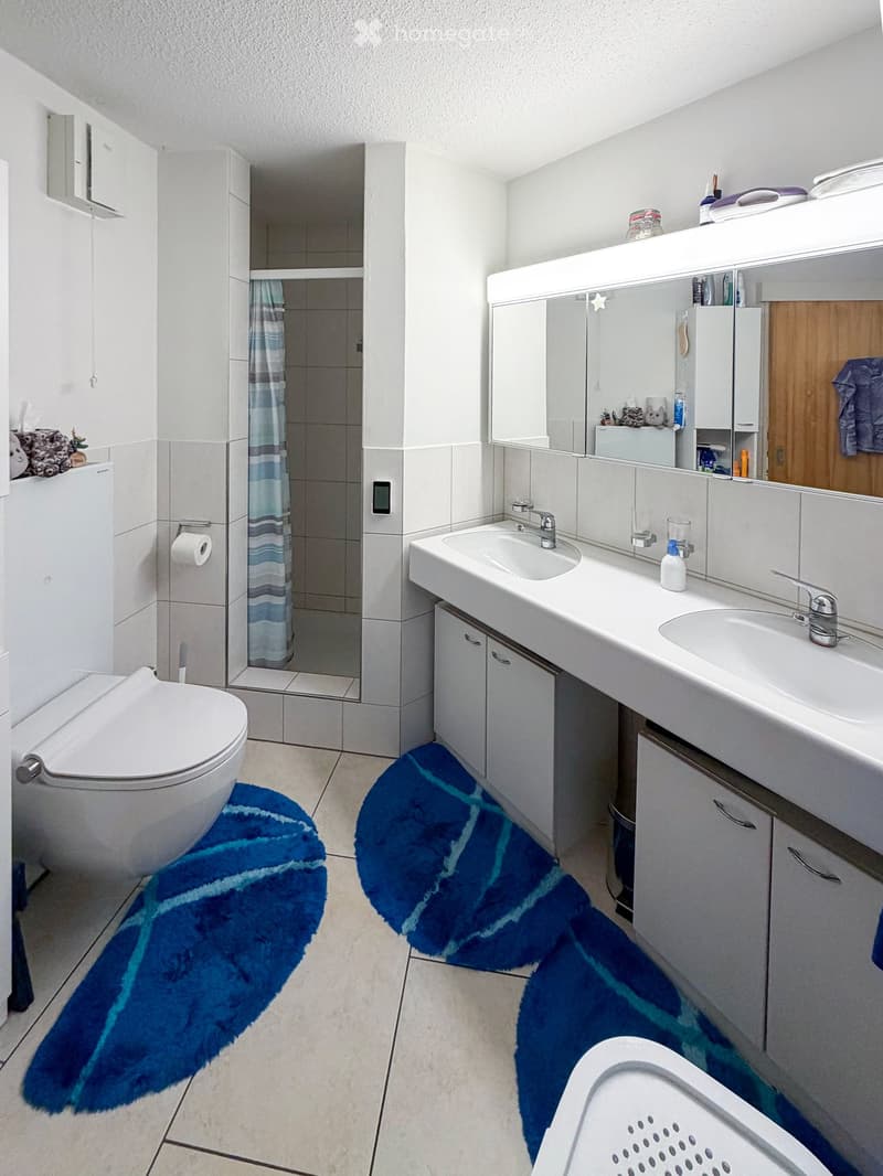 Modern bathroom with double sink, toilet, large mirror, and shower area with a blue and white striped shower curtain. Blue bath mats are placed in front of the toilet and sink.