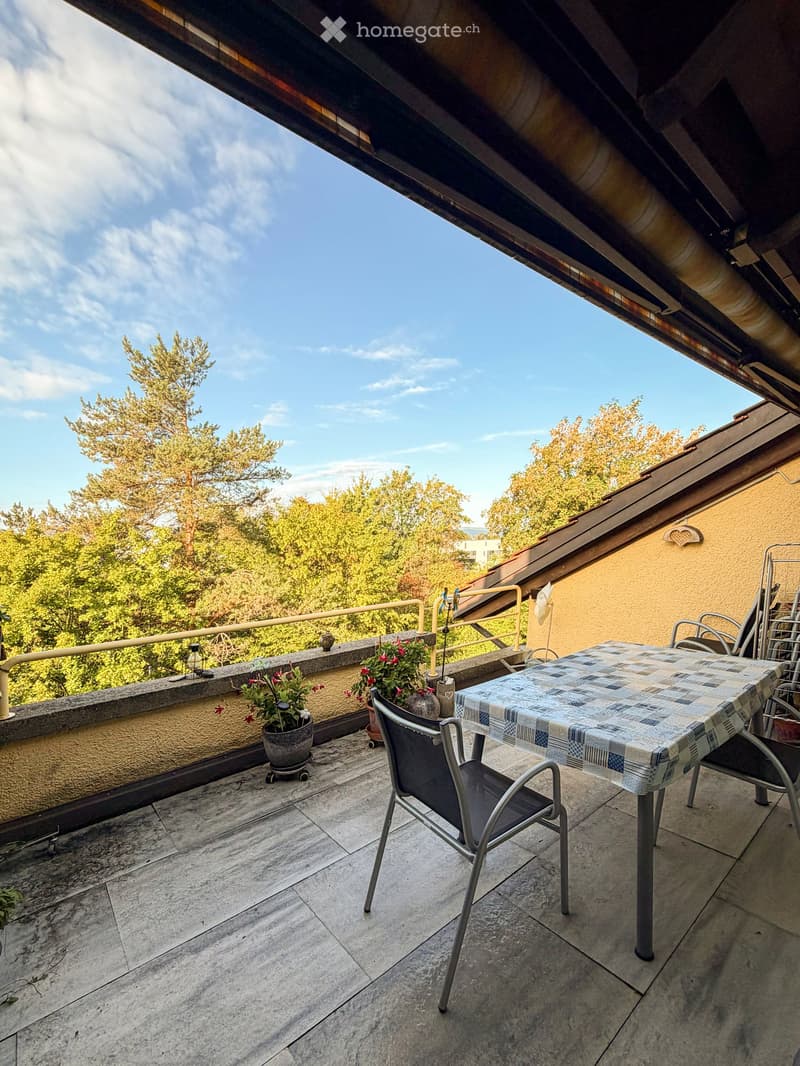 Balcony with a table, chairs, and potted plants, overlooking a tree-filled area.