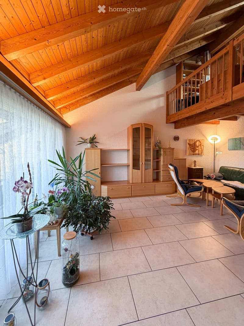 Bright living room with wooden beams, large windows, and a staircase leading to an upper level. Features a sofa, chairs, a coffee table, a wooden cabinet, and several potted plants.