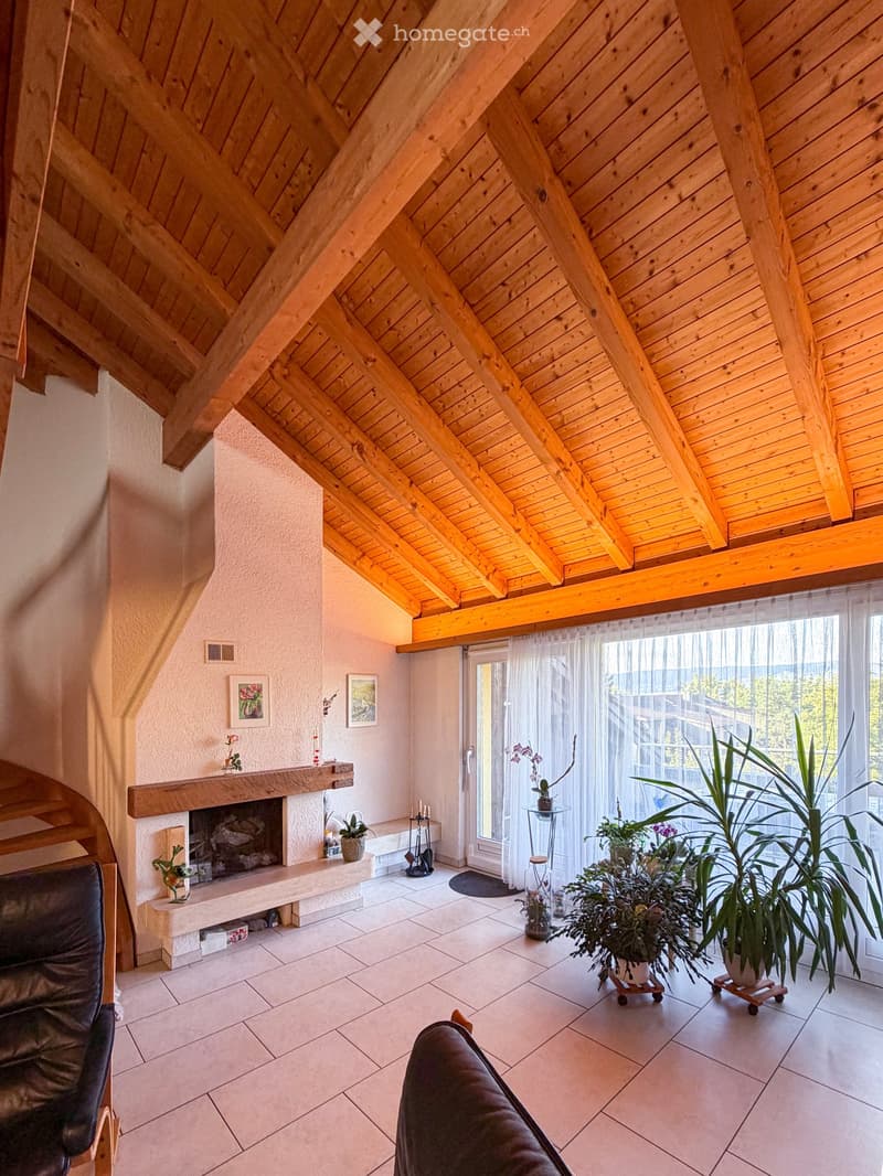 Living room with exposed wooden beams, fireplace, large window with view, sofa, and plants.