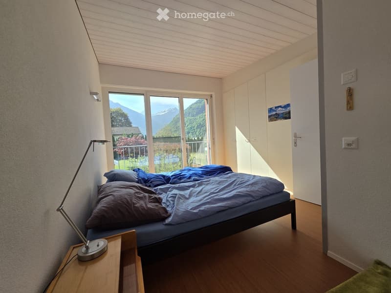 Cozy bedroom with wooden flooring, a bed with blue bedding, a nightstand with a lamp, and large windows offering a scenic mountain view.