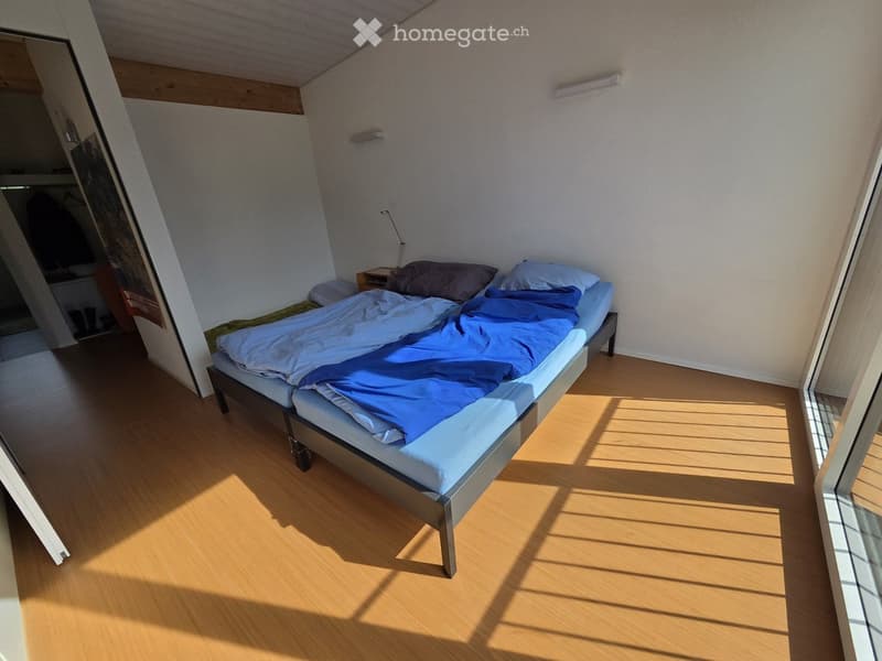 A modern, minimalist bedroom with a single bed, nightstand, and lamp. The room features light wooden flooring and white walls, with natural light streaming in from a large window.