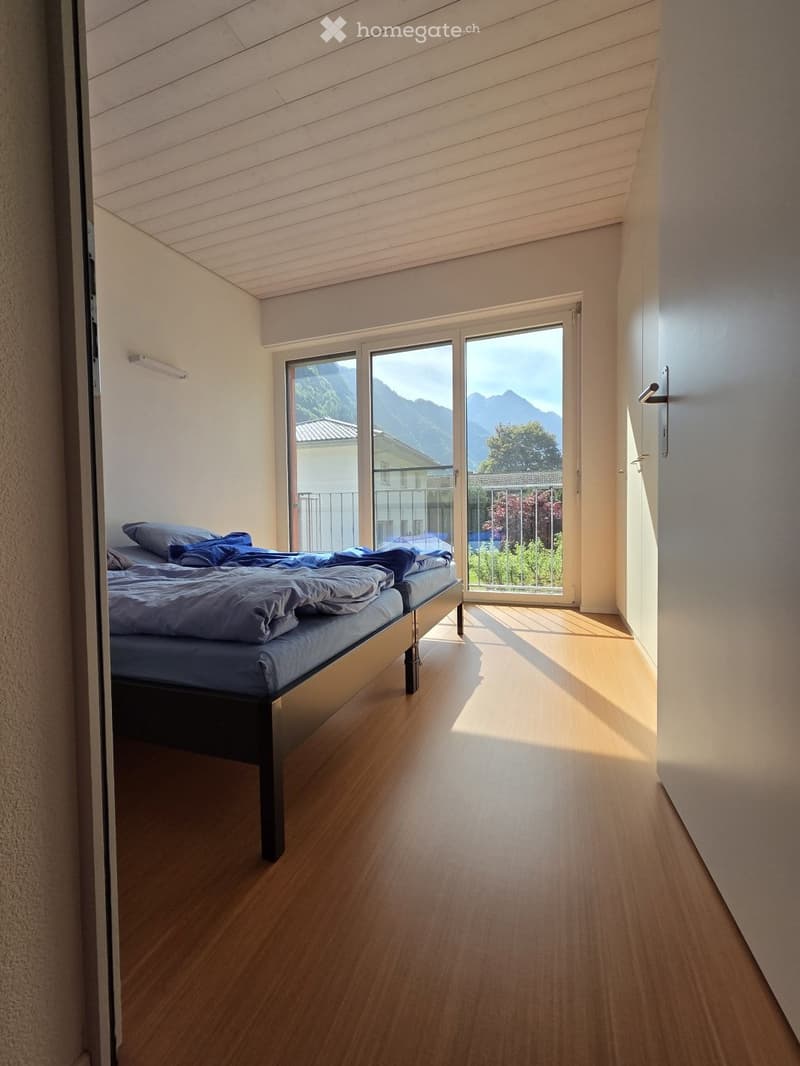 Bright bedroom with wooden flooring, a single bed with blue bedding, large windows offering a scenic mountain view, and modern interior design.