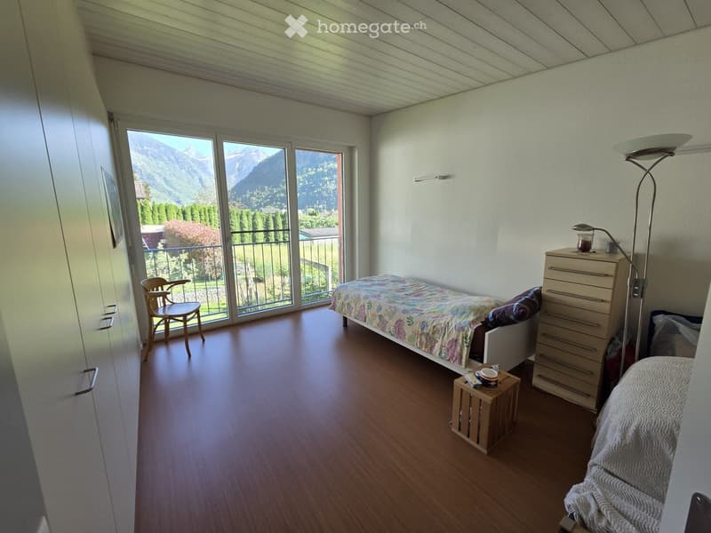 Bright bedroom with wooden flooring, large window offering a scenic mountain view, bed with colorful bedding, nightstand, chair, and a tall floor lamp.