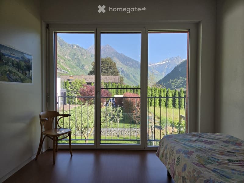 Bright bedroom with large windows offering a scenic mountain view, featuring a bed with a colorful bedspread and a wooden chair near the window.