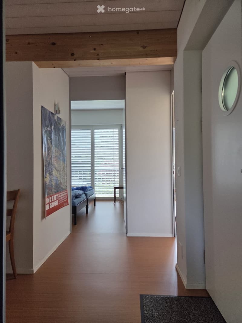 A modern hallway with wooden flooring, white walls, and a large circular window on the right side. A poster is displayed on the left wall, and a small table and chair are visible in the adjacent room.