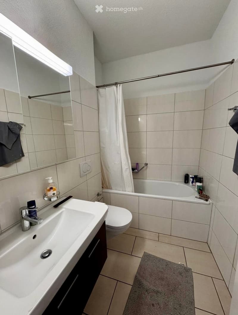 Modern bathroom with a white sink, toilet, bathtub, and mirror. The space features tiled walls and floor, a shower curtain, and a towel on the side.