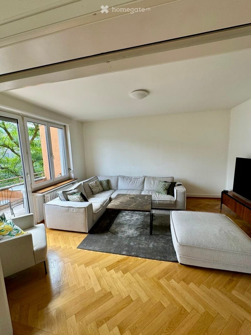 Modern living room with light-colored sectional sofa, coffee table, ottoman, armchair, and television. The room features wooden flooring and large windows with a view of greenery.