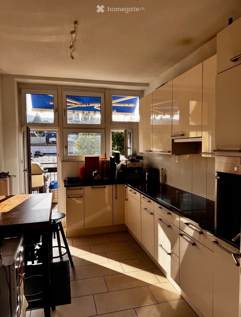 Modern kitchen with light-colored cabinets, black countertops, and a central island with a stool. Appliances include an oven and coffee maker, with a window offering a view of the outside.