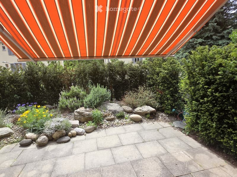 Garden area with a stone-paved patio, large rocks, flowering plants, and a retractable awning providing shade.