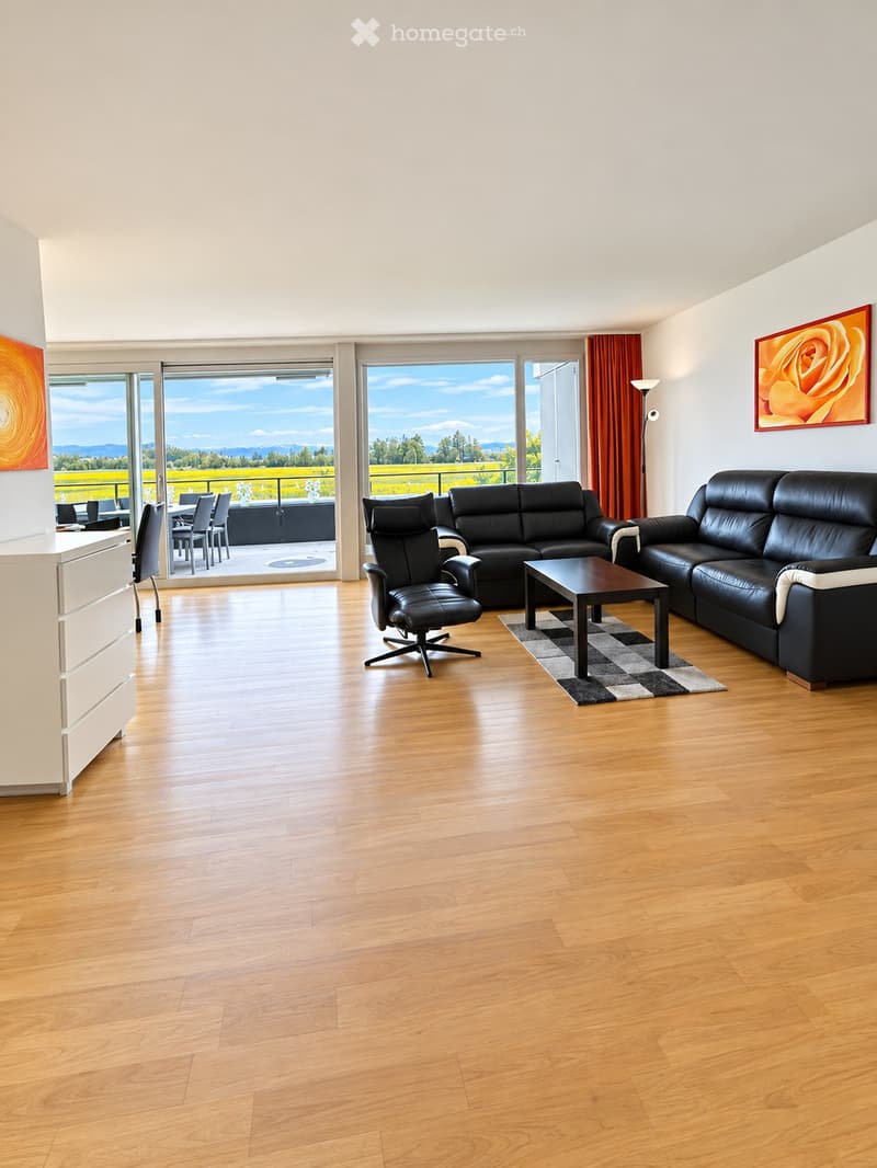 Modern living room with hardwood floors, large windows offering a scenic view, black leather sofa, coffee table, and a dining area visible through the open sliding doors.