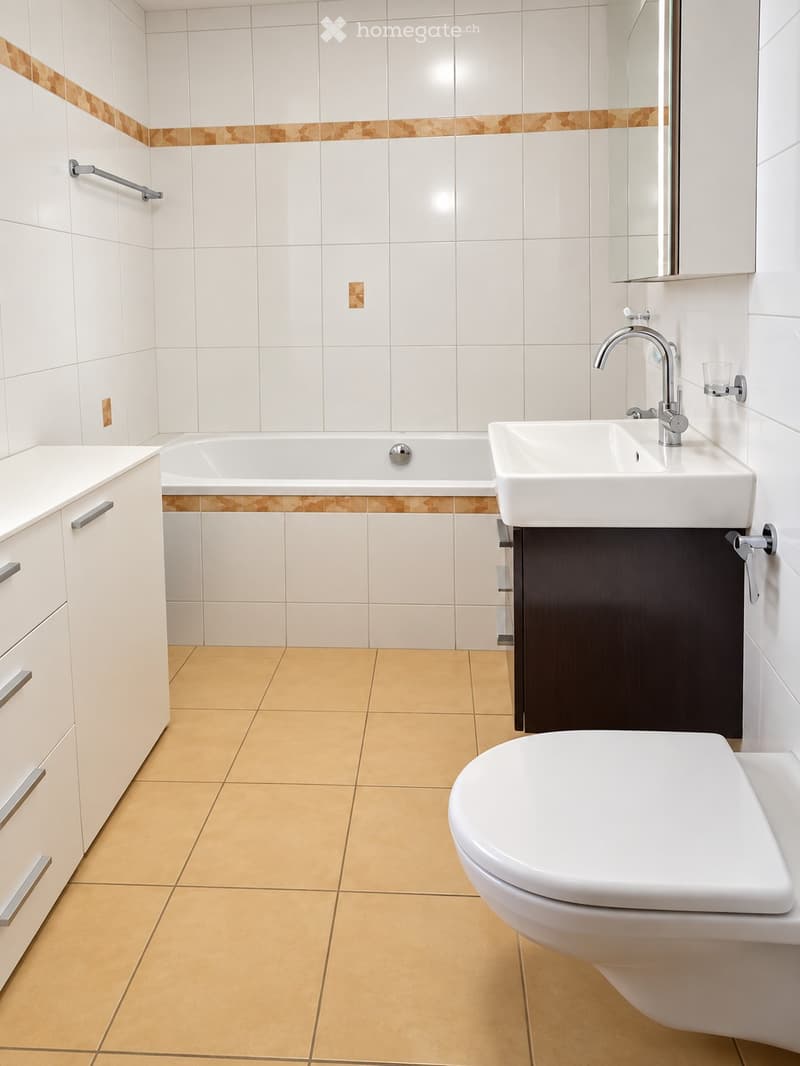 Modern bathroom with white tiles, beige floor tiles, a bathtub, a sink, a toilet, and a mirrored cabinet.