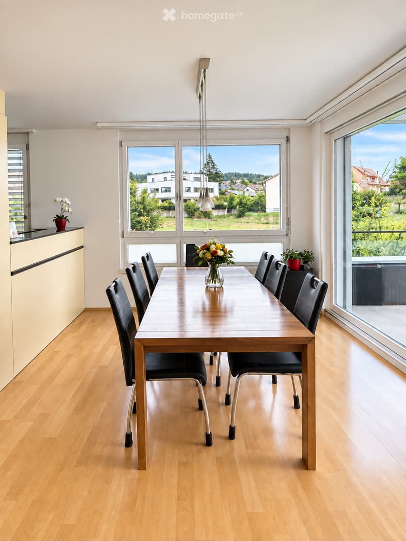 Bright dining room with wooden floor, large windows, a long wooden dining table surrounded by black chairs, a vase with flowers in the center, and a potted plant on the windowsill.