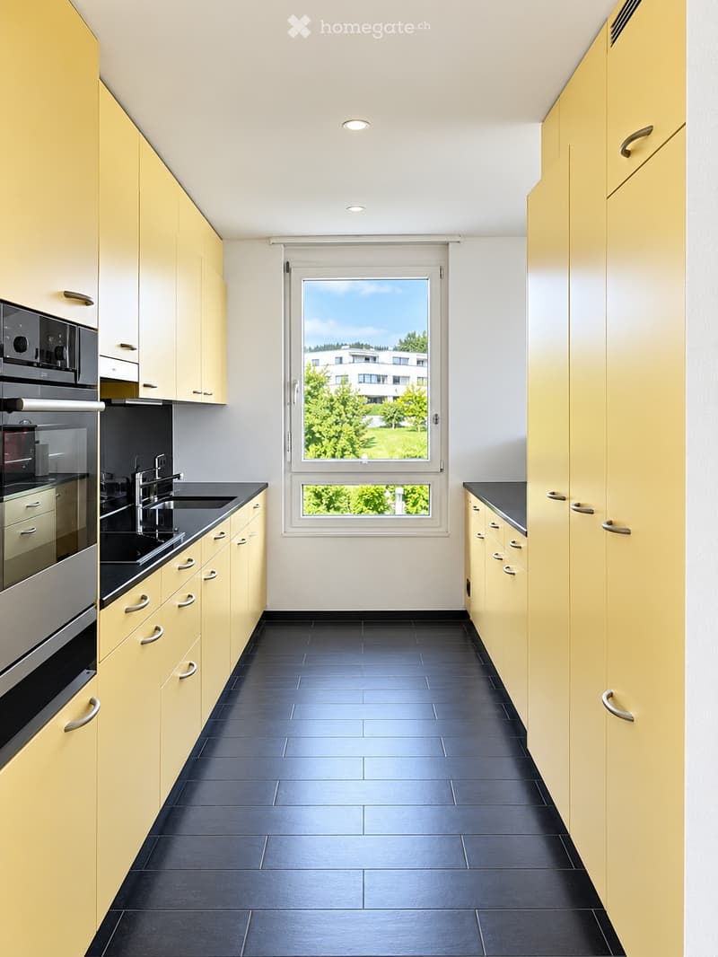 Modern kitchen with light yellow cabinets, black countertops, and dark tiled flooring. Features a window with a view of greenery and a building outside.