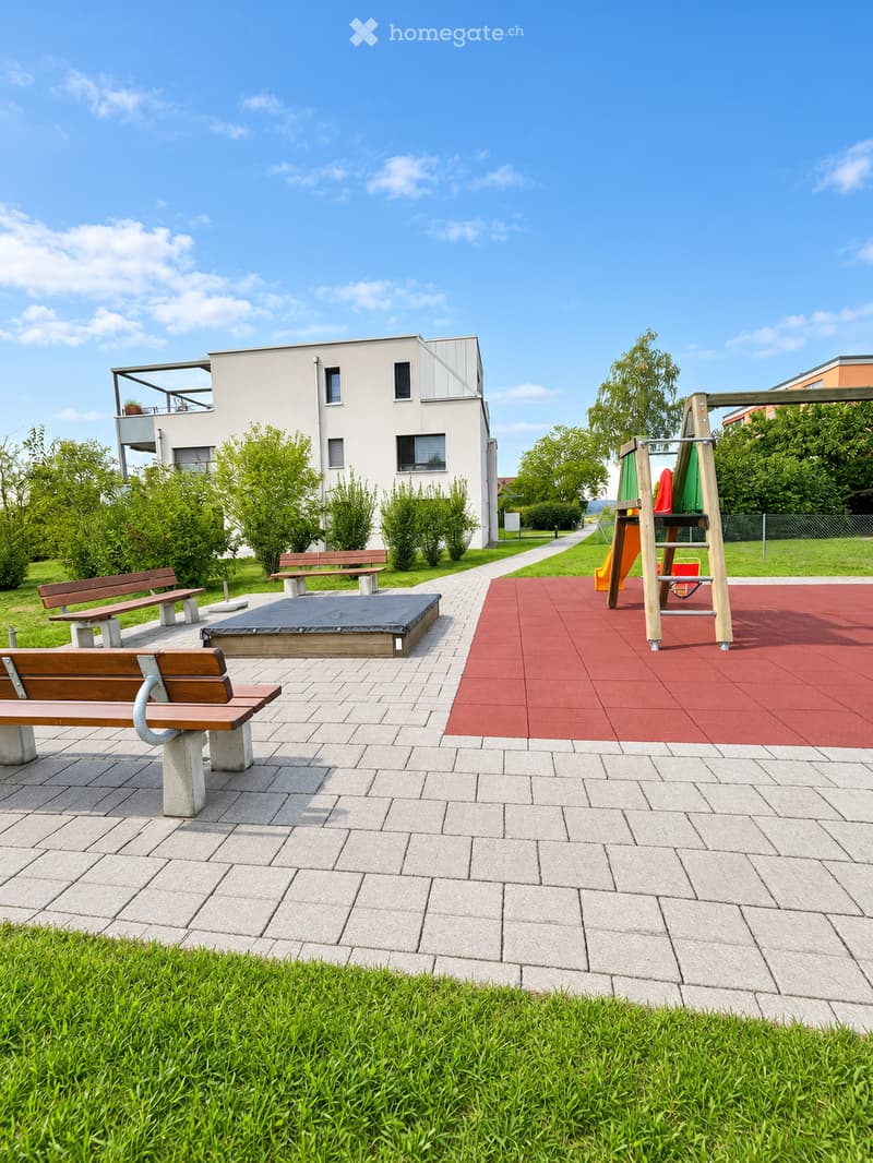 Outdoor area with benches, playground equipment, and a modern building in the background.