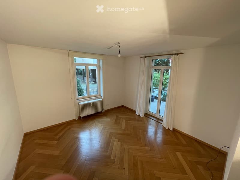 Empty room with wooden herringbone flooring, white walls, large windows with white curtains, radiator, and a ceiling light fixture.