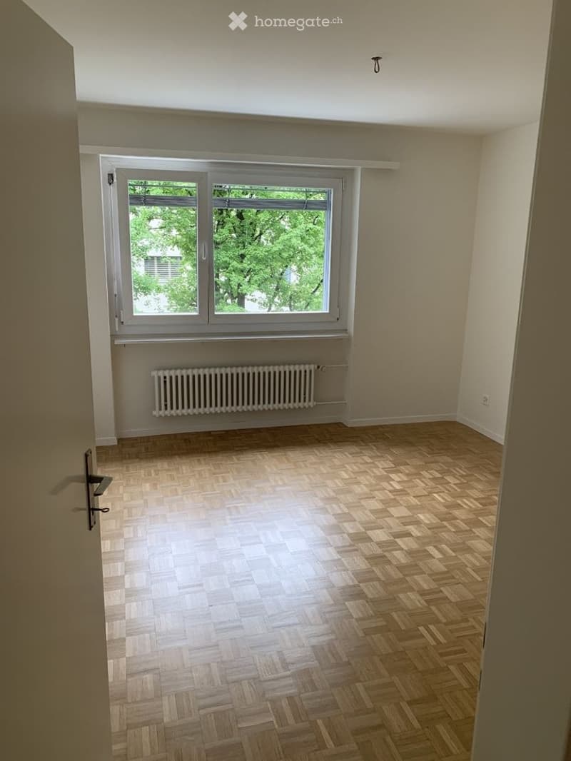 Empty room with wooden parquet flooring, white walls, a large window with white frame, and a radiator below the window.