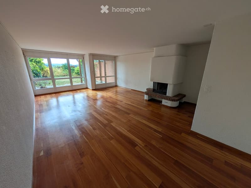 Empty room with wooden flooring, large windows, and a fireplace with a bench.