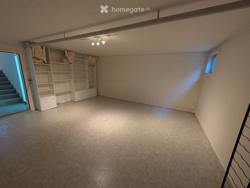 Empty basement room with concrete floor, white brick walls, built-in shelving unit, small window, and staircase leading upwards.