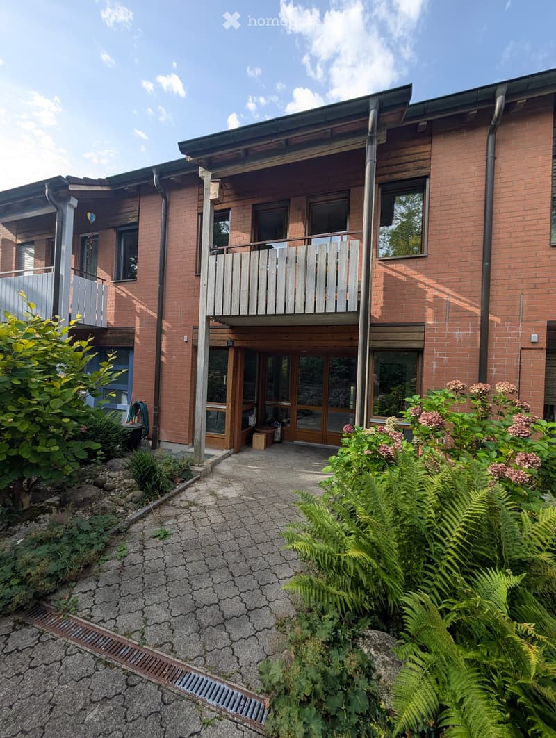 Two-story brick building with a balcony, surrounded by greenery and a paved pathway.