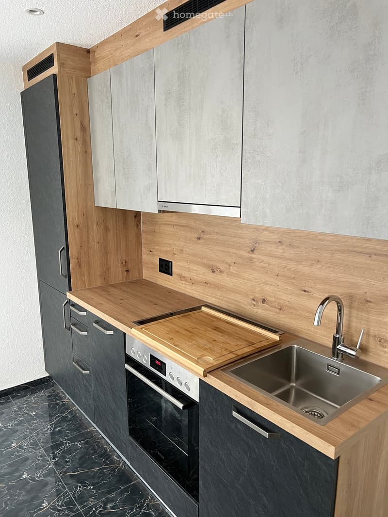 Wooden cabinets, gray stone walls, white granite countertop, wooden chopping board, stainless steel sink, silver faucet, black marble flooring, modern stove and oven, power outlet