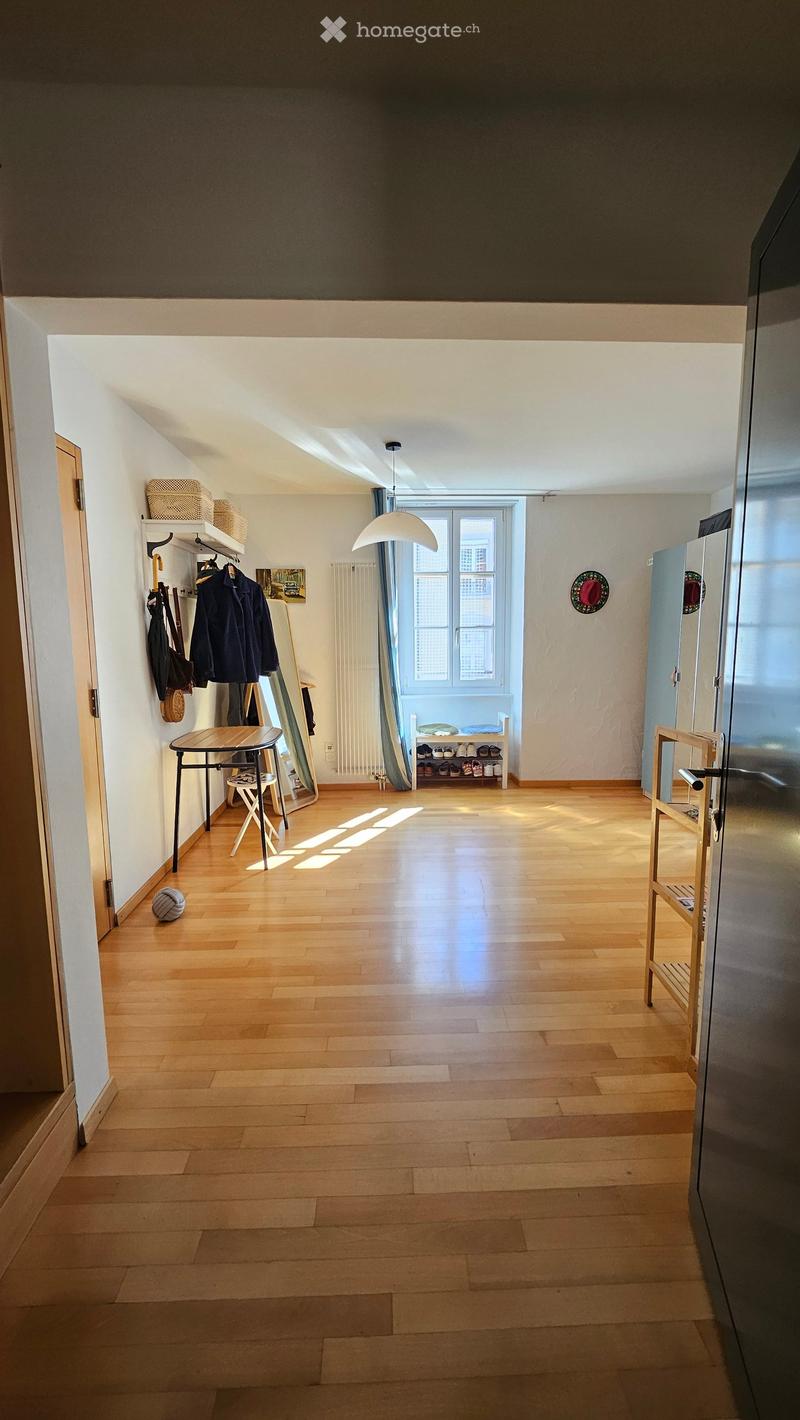 Bright hallway with wooden flooring, white walls, and natural light streaming in from a window at the end. Features include a coat rack with jackets and bags, a small table with a chair, a shoe rack, and a shelf with baskets and a mirror.
